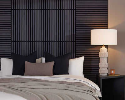Bedroom with black wall panelling, a white bed, black and brown cushions, a beige throw, black side table and a stone-effect lamp.