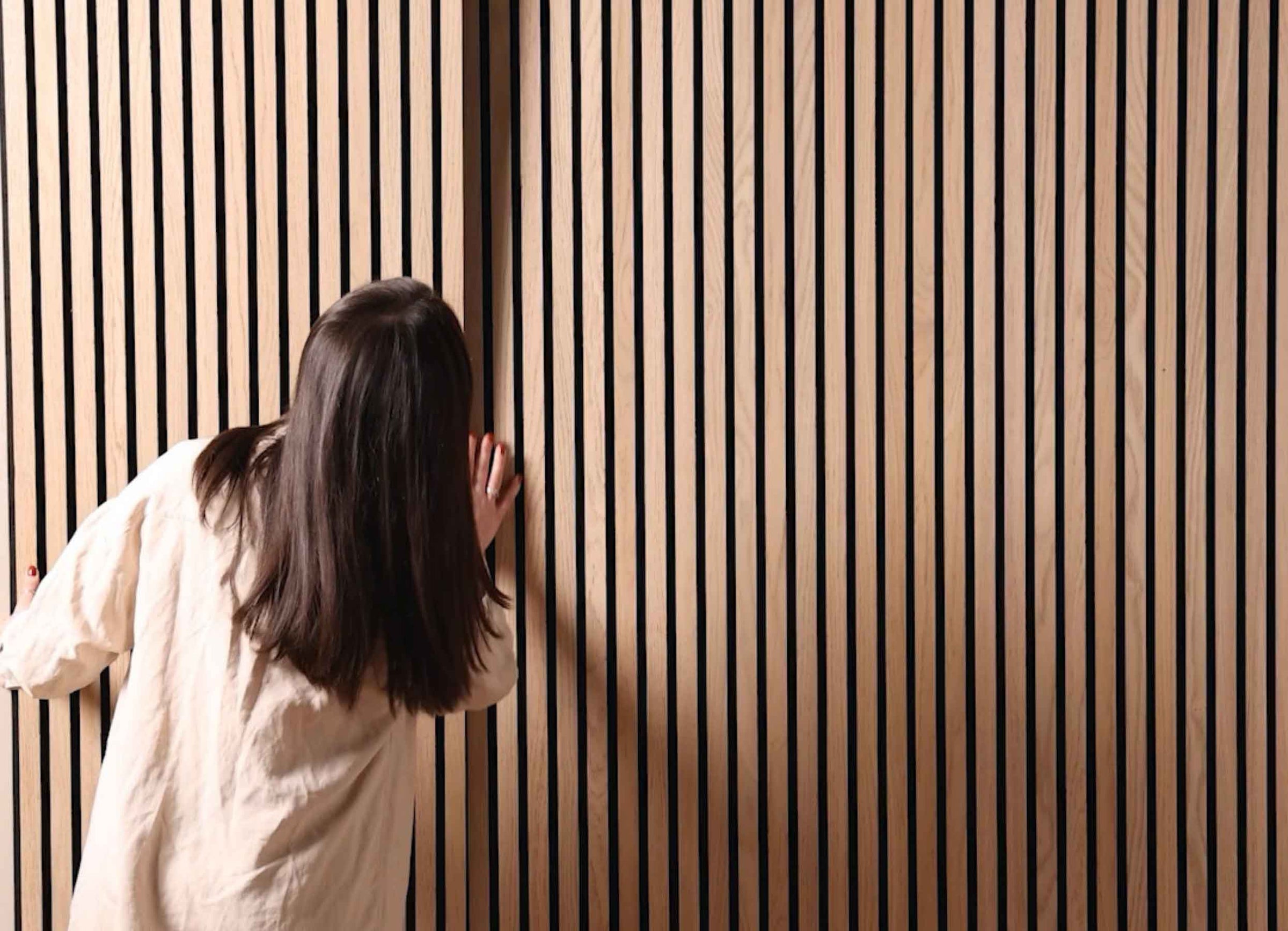 Dark-haired woman wearing a beige shirt placing a SlatWall Natural Oak panel on a wall.