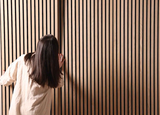 Dark-haired woman wearing a beige shirt placing a SlatWall Natural Oak panel on a wall.