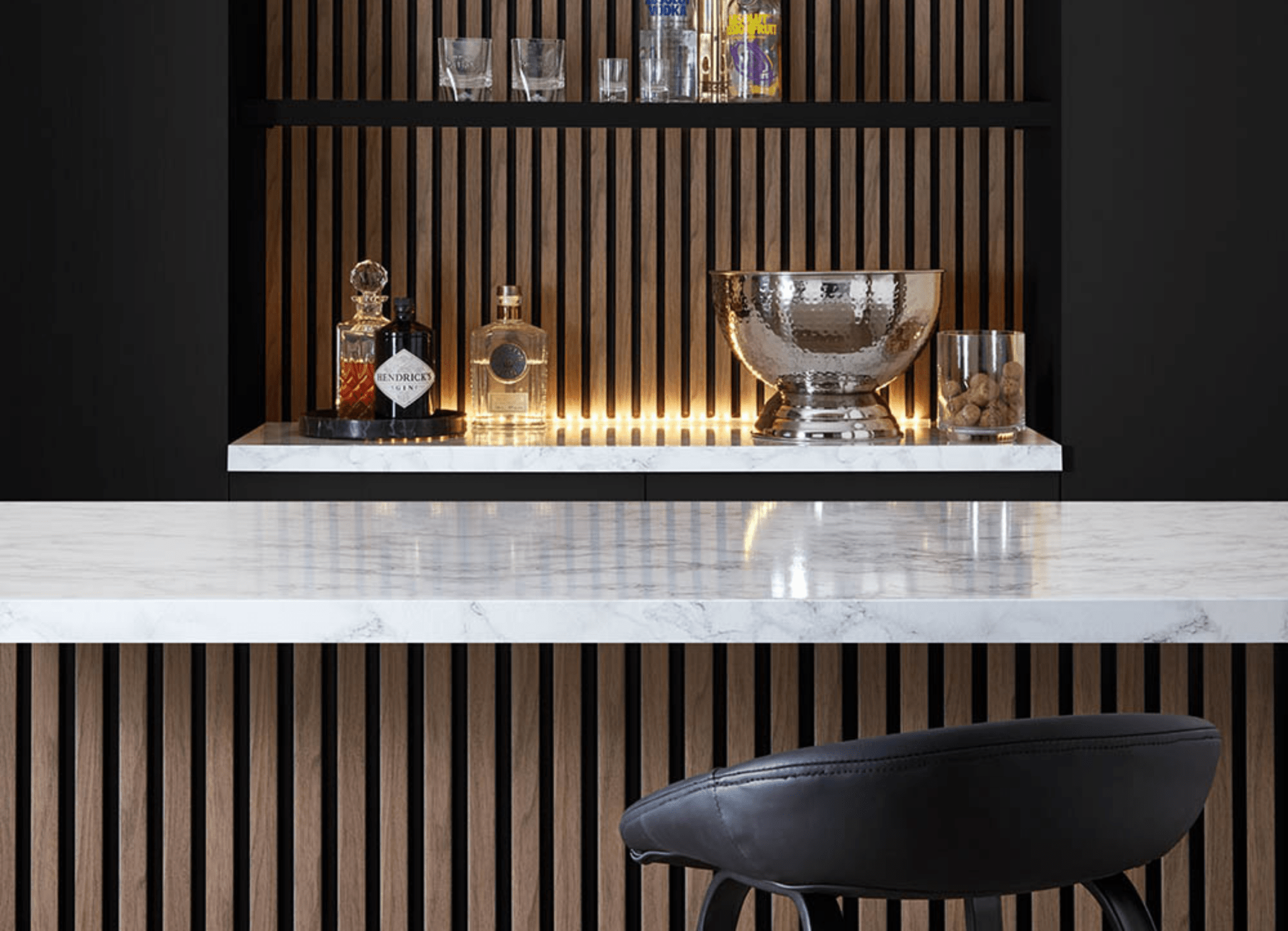 This image shows a kitchen island with the SlatWall behind the shelves and a bar stool. The shelves display bottles of alcohol and an ice bucket 