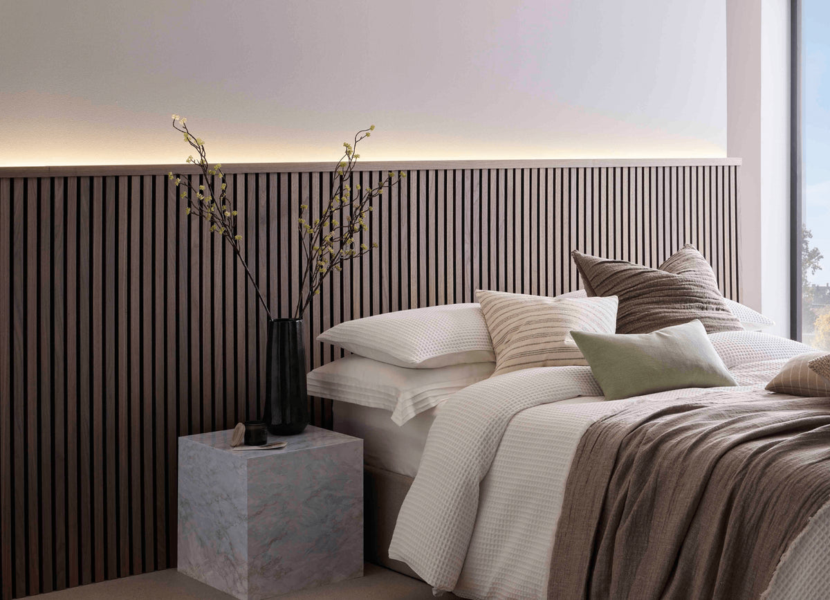 SlatWall Midi Walnut panels and coordinating Top Trim behind a marble side table and a white bed with various cushions.