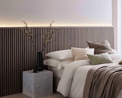 SlatWall Midi Walnut panels and coordinating Top Trim behind a marble side table and a white bed with various cushions.