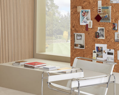 A cork pinboard featuring pictures behind a row of books and a white and silver chair, next to a window and SlatWall panels.