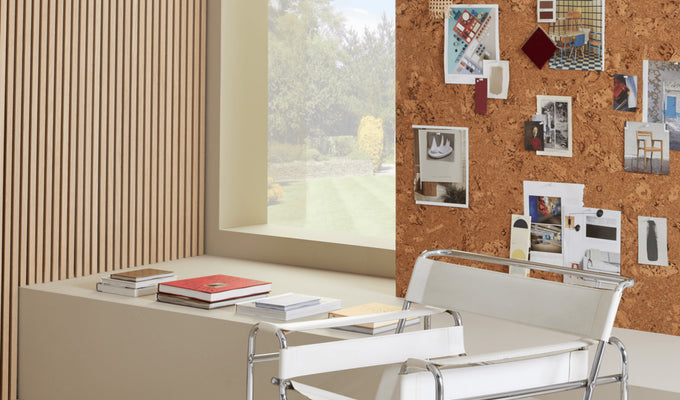 A cork pinboard featuring pictures behind a row of books and a white and silver chair, next to a window and SlatWall panels.