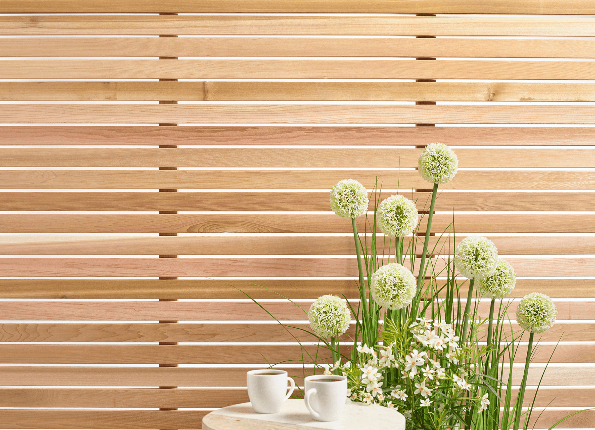 Cedar wood outdoor panels behind green and white flowers next to a table with two coffee cups.