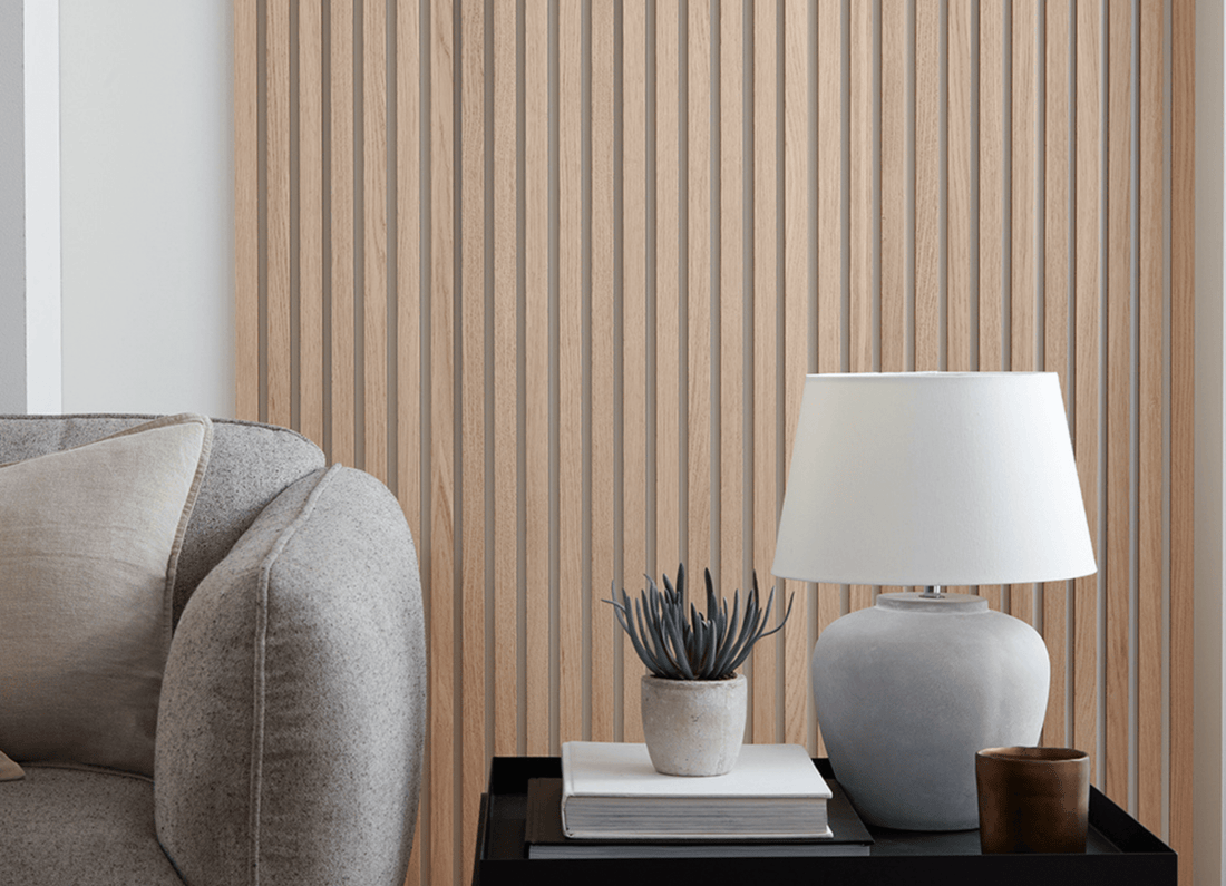 SlatWall Natural Oak individual wooden wall slats behind a grey sofa and black table with books, a plant and a lamp on top.