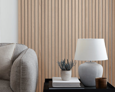 SlatWall Natural Oak individual wooden wall slats behind a grey sofa and black table with books, a plant and a lamp on top.