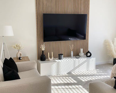 Light interior space with homely decor. Including a cream sofa, with black cushions, and a Naturewall SlatWall feature on the wall surrounding a TV.